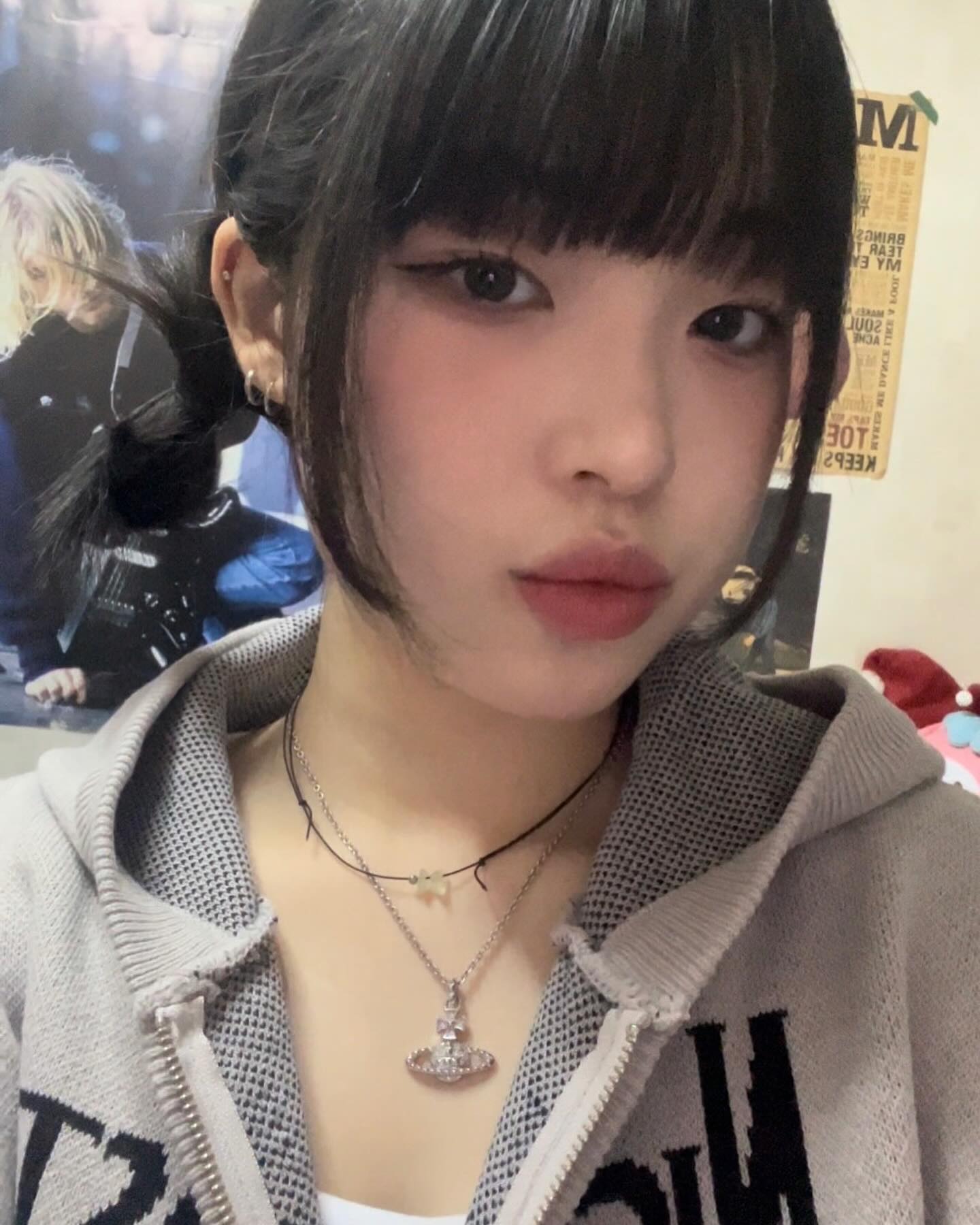 a young woman with black hair in a space buns hairstyle takes a selfie. she is wearing a grey zipup hoodie with a white graphic, and three necklaces  one with a small charm, one with a small cross, and one with an angel pendant. she has simple makeup with winged eyeliner. she is standing in front of a poster. the image is welllit.