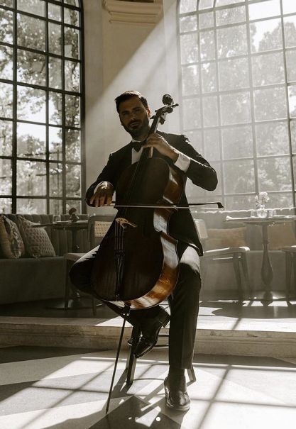 a man in a black suit sits elegantly in a sunlit room, playing a cello. large windows with dark frames illuminate the space, casting shadows and highlighting the architectural details. the overall atmosphere is sophisticated and artistic.