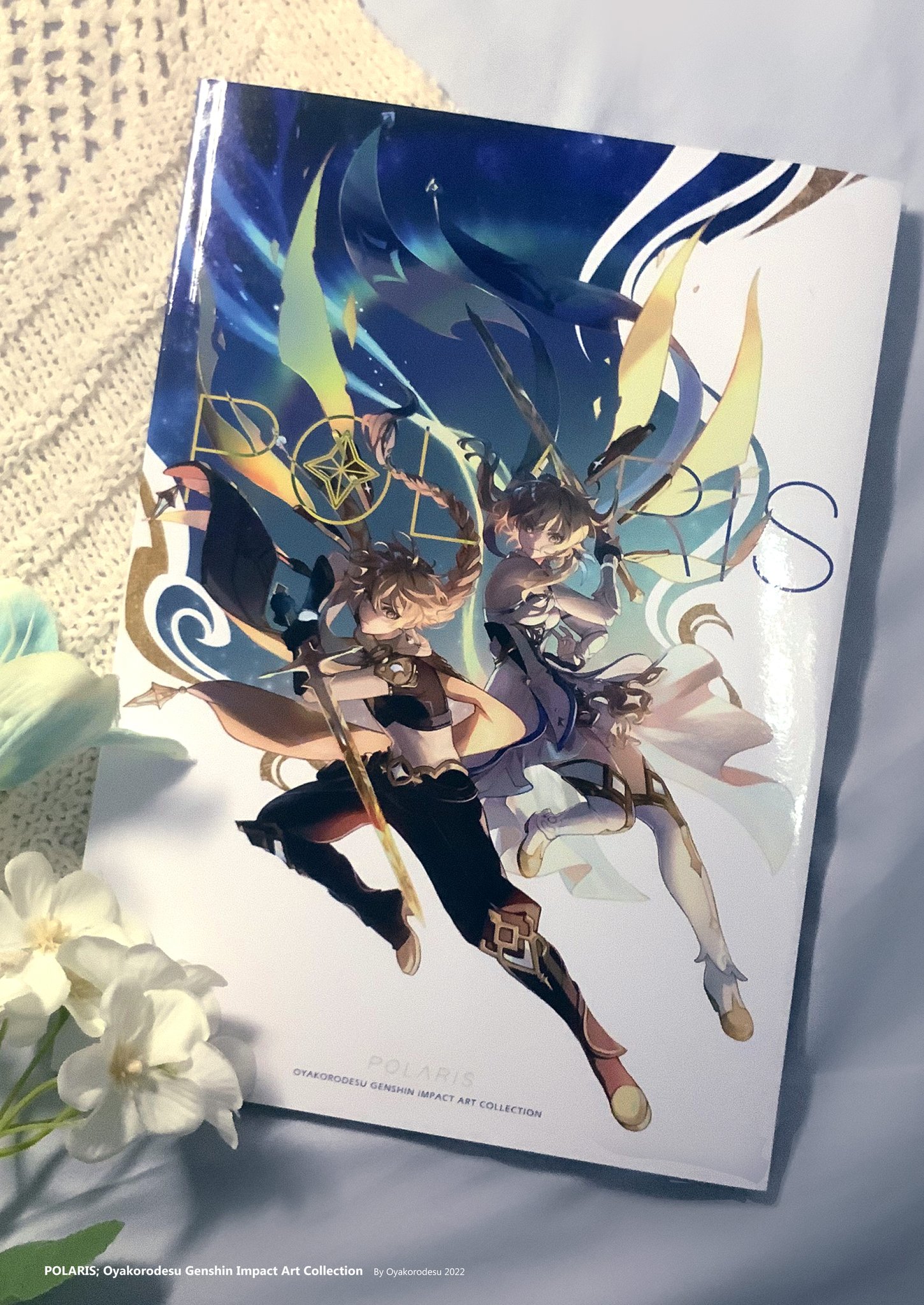this is an image of the cover of an art book for the popular video game genshin impact. the book is titled "polaris: oyakorodesu genshin impact art collection." the cover art features two characters from the game, aether and lumine. they are both depicted in their signature outfits, aether in his explorer garb and lumine in her dress, with a white and blue background, featuring what appears to be a starry sky. the book is lying flat on a white knitted surface. next to the book are white flowers, some of which are out of focus.