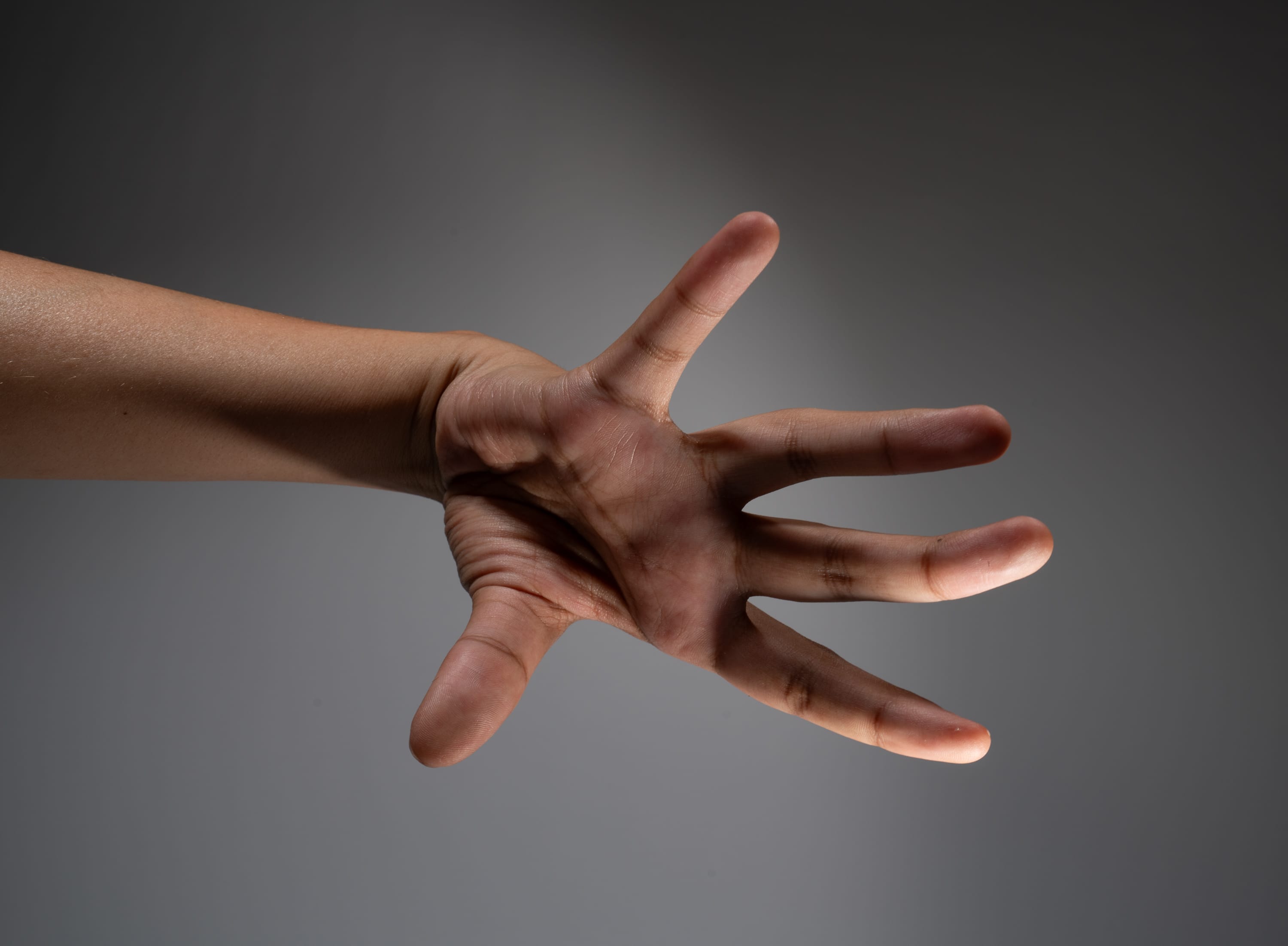 the image showcases a single hand extended against a neutral gray background. the hand is reaching forward with an open palm and slightly spread fingers. the lighting emphasizes the contours and texture of the hand, creating a dramatic and compelling composition.
