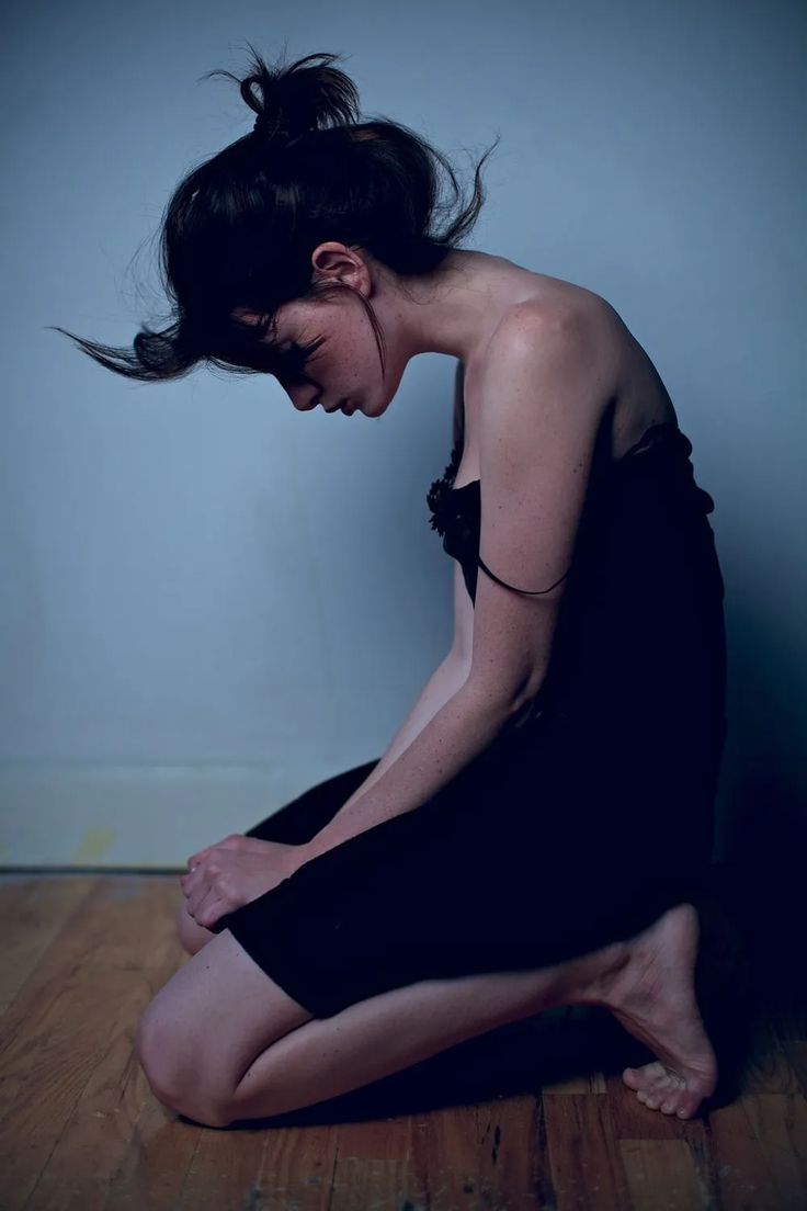 a young woman with long, dark hair is sitting on a wooden floor. she is wearing a black dress and her hair is styled in a messy bun. she is looking down and appears to be sad. the image is softfocused and has a dramatic feel.