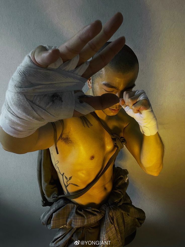 a man is shown shirtless in a fighting stance with his left hand raised in front of his face as if to defend a blow.  his right hand is held near his face, partially obscured by his left arm, in a fist. he has black ink tattoos on his chest and side. the lighting in the photograph is dark, with some bright, yellow light coming from the bottom and right side of the frame, almost illuminating his bare torso. the image gives a sense of strength and athleticism.