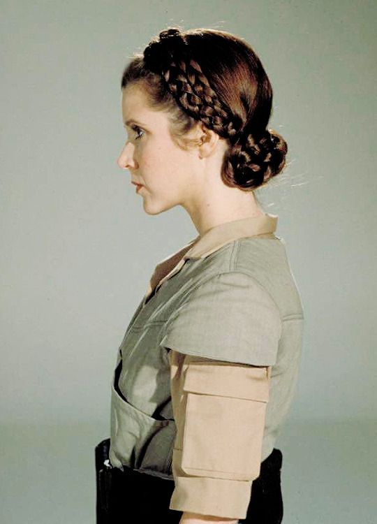 a profile view of carrie fisher as princess leia from the 1977 movie "star wars: a new hope," with her signature braided hair and a beige and green costume.