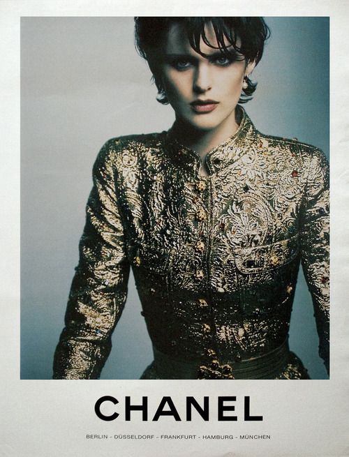 a vintage chanel advertisement features a closeup portrait of a woman model. she's wearing a richly detailed gold brocade jacket, with an elegant, sophisticated style characteristic of the 1990s.  the background is a muted grey, allowing the model and her outfit to stand out. the chanel logo is prominently displayed at the bottom, along with a list of german cities: berlin, düsseldorf, frankfurt, hamburg, münchen.  the overall mood is one of high fashion and timeless elegance.
