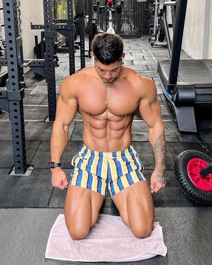 a muscular man with a welldefined sixpack is kneeling on a towel in a gym. he's wearing yellow and blue striped shorts and a gold necklace.  he appears to have earphones in his ears. the background shows various gym equipment, including weights and a treadmill. the man's physique is very athletic and muscular, suggesting a dedicated fitness routine. he has tattoos on his arms.