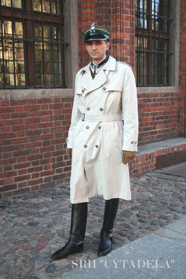 a man in a military uniform and trench coat stands in front of a brick building. he is wearing a hat and boots. the ground is cobblestone.