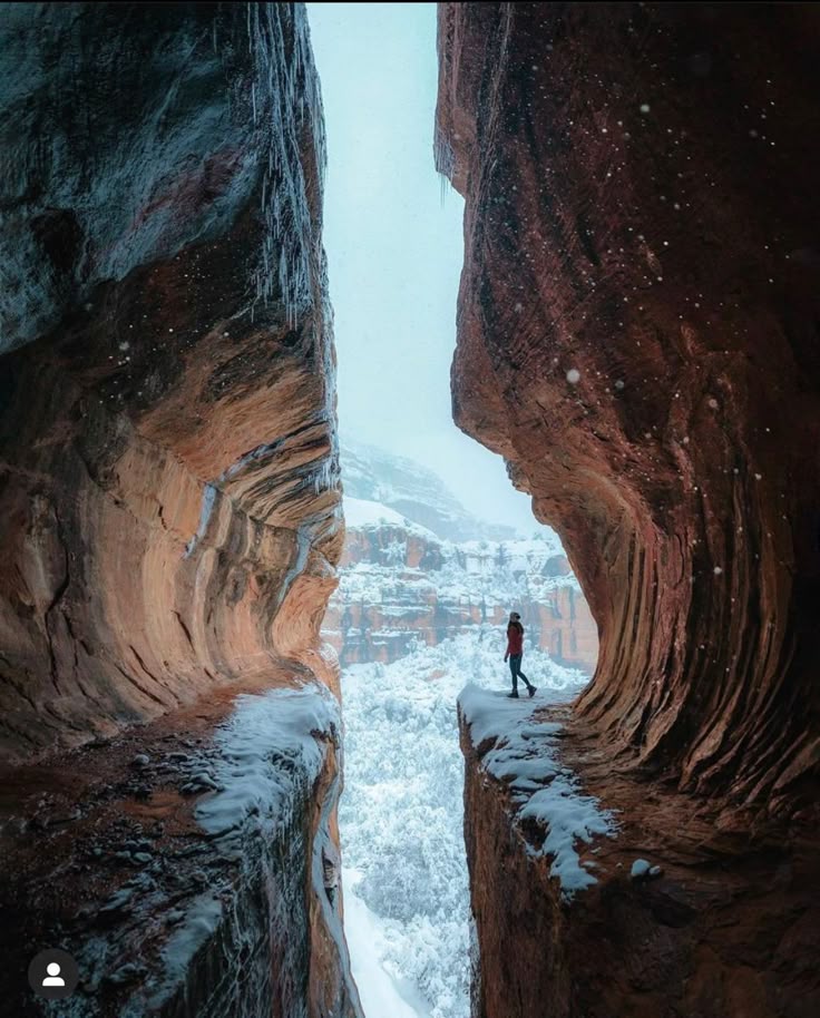 a person stands on the edge of a cliff between two canyon walls covered in snow. the background shows a snowy mountain range. the scene is wintery and adventurous.