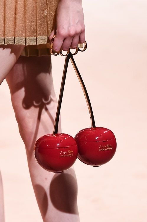 a person is holding a red cherryshaped handbag with a gold handle. the handle has a ringlike design, and the person is wearing a gold dress. the background is a pale peach color.