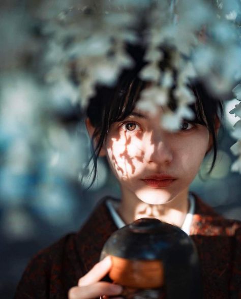 a young woman with black hair is looking at the camera while holding a teacup. she's wearing a red kimono and surrounded by cherry blossoms.
