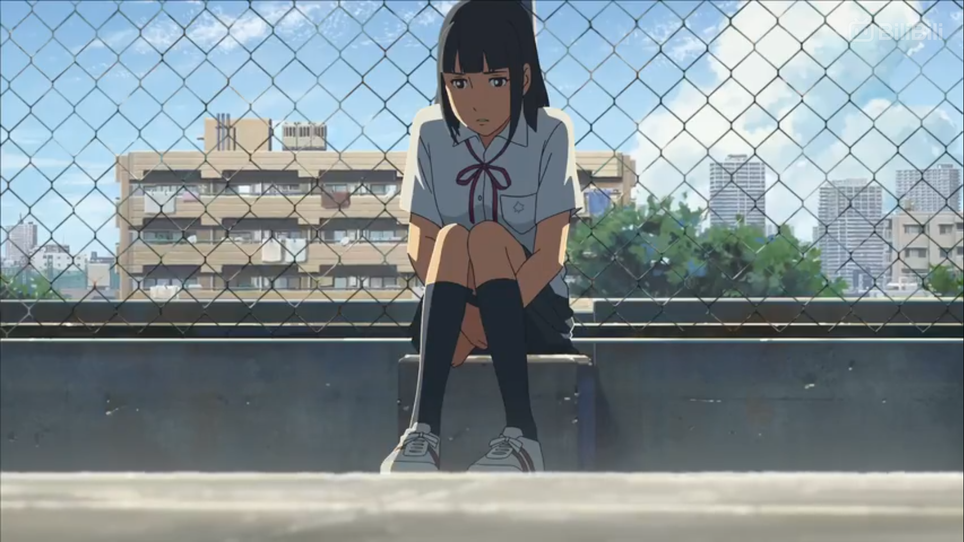 the image shows a young anime girl with short black hair and a sad expression sitting alone on a rooftop. she is wearing a typical japanese school uniform. a chainlink fence separates her from the backdrop of a sunny cityscape with buildings and blue skies.  the overall atmosphere conveys a sense of loneliness and contemplation.