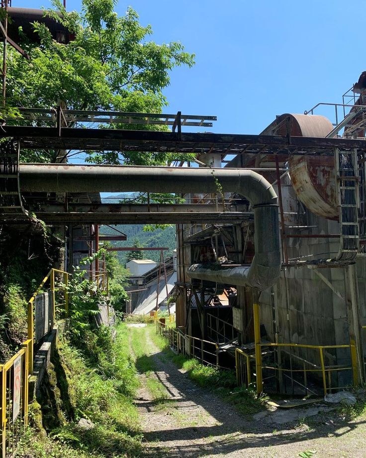 the image shows a pathway leading through the overgrown ruins of an abandoned industrial complex, possibly a factory or mill.  the structures are heavily rusted and decaying, with large pipes and metal frameworks visible.  lush green vegetation has taken over much of the site.  the background shows distant hills or mountains, suggesting a rural setting. the sunlight and shadows indicate it's daytime in summer. the overall impression is one of decay and abandonment amidst a reclaiming natural environment.