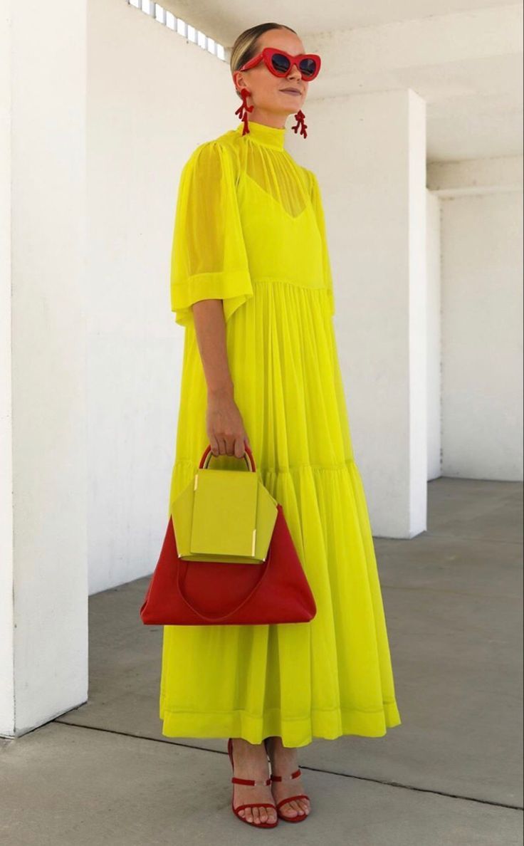a woman is wearing a bright yellow maxi dress with a high neck and 3/4 sleeves. she is also wearing red sunglasses, red earrings, and red high heeled sandals. the woman is holding a red bag with a yellow top in her hands. the woman looks stylish and elegant, and she is ready to enjoy the summer. she is standing in front of a white wall.