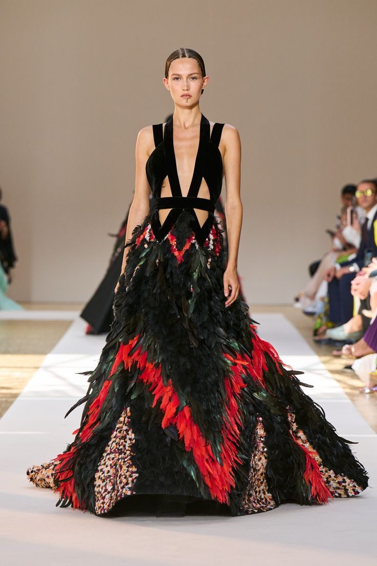 a fashion model walks the runway in a stunning black dress adorned with red and green feathers. the dress has a daring, lowcut neckline and is cinched at the waist with a belt. the model's hair is pulled back, highlighting her elegant features. she poses confidently, radiating beauty and glamour. the dress is a striking example of haute couture design, showcasing intricate details and exquisite craftsmanship. the backdrop of the runway creates a dramatic and luxurious setting for this fashion show.