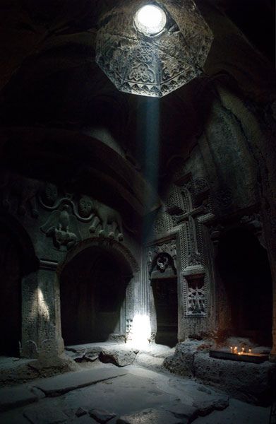 the interior of a cave church, possibly within the gegard monastery in armenia, featuring intricate stone carvings, including armenian crosses and archways. light streams in through a skylight, illuminating the dark, cavernous space. candles are lit near a wall, adding to the ambiance.