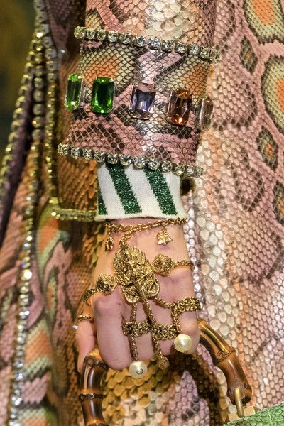 loseup shot of a model's hand adorned with elaborate gold jewelry, including intricately designed rings and a bracelet, set against a background of a snakeskin patterned gucci garment. the jewelry features various gemstones and pearls, adding to its luxurious and opulent appearance. the focus is on the detail and craftsmanship of the accessories, emphasizing the rich textures and patterns. the image is indicative of high fashion and luxury, highlighting gucci's signature style.