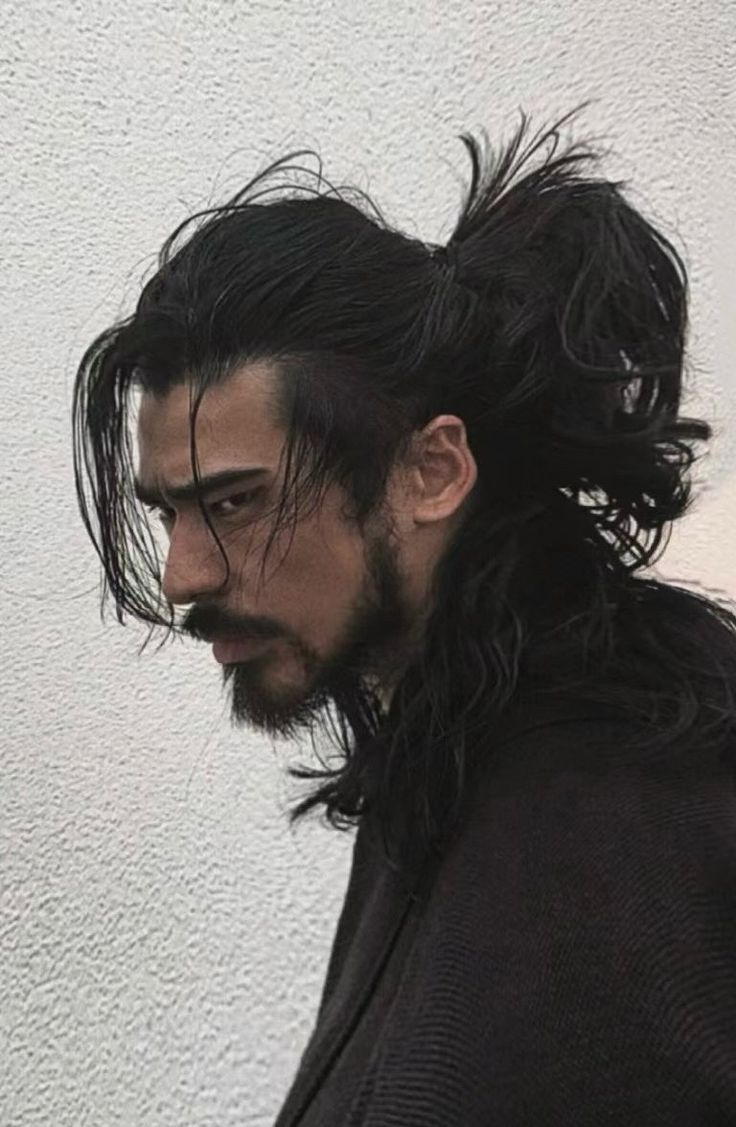 a profile shot of a man with long, dark hair pulled back into a high ponytail. he has a beard and mustache. he is wearing black clothing, and the background is a textured white wall.