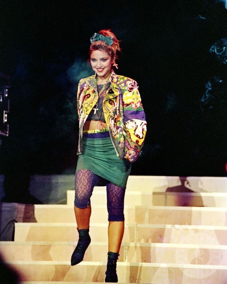 madonna is shown in a full shot descending stairs on a stage, during a performance. she is wearing an ensemble that includes a colorful patterned jacket, a matching short skirt, caprilength fishnet leggings, and black ankle boots. her hair is styled with a green bow. she is smiling as she walks down the stairs. the background is dark, with stage lighting and some smoke effects visible.