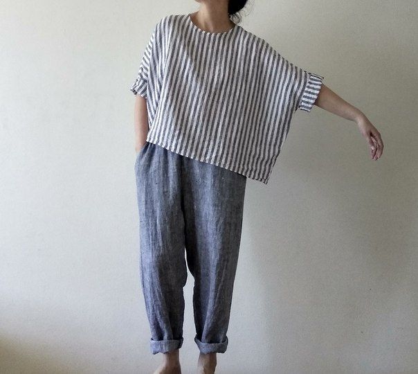 a woman stands against a plain background, wearing a loosefitting, striped top with short sleeves and a pair of grayblue linen pants with cuffed bottoms. the outfit suggests a casual and comfortable style.