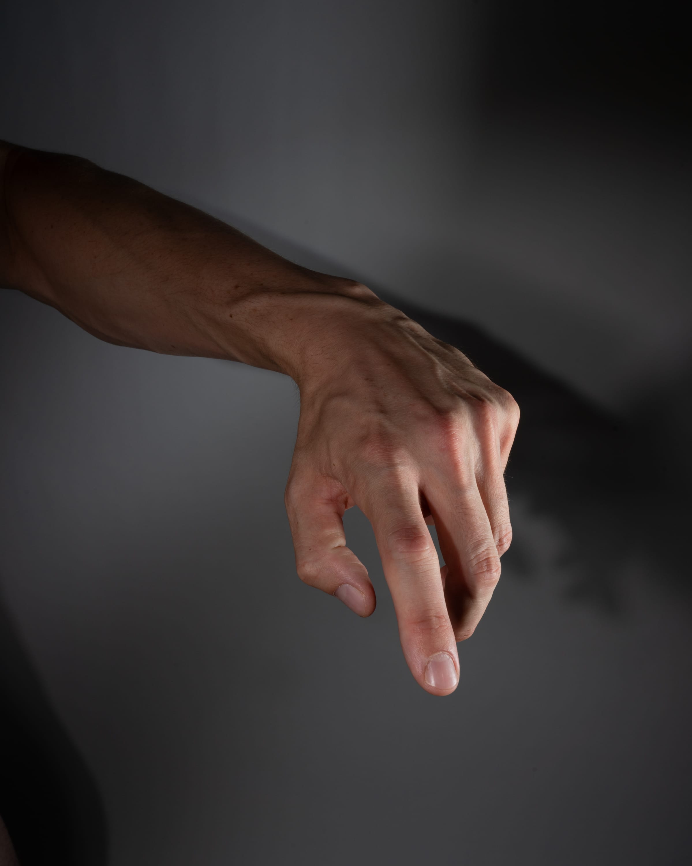 a man's arm and hand reaching down, against a gray background. the hand is partially open, with the fingers slightly curled. the veins in the arm and hand are visible. the lighting is soft and diffused, creating subtle shadows.