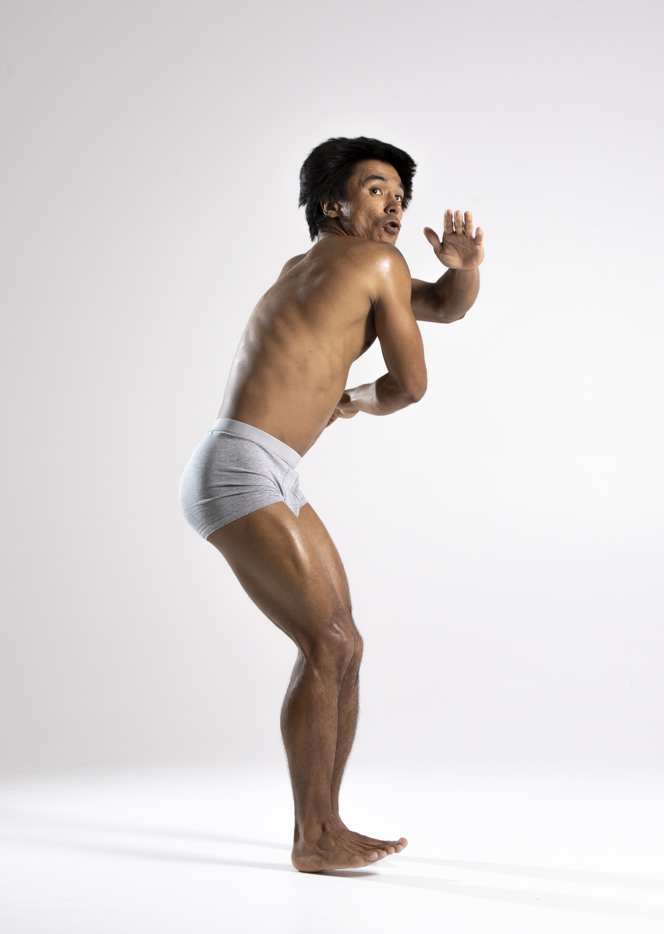 a shirtless man wearing gray underwear is standing on a white background. he has a scared expression on his face and is looking back over his shoulder with his mouth open. he has one hand raised in front of him as if to ward off something. the lighting is bright and even, highlighting his muscular build. his body appears to be oiled.
