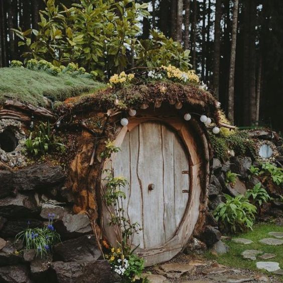 an enchanting hobbit house nestled amidst a lush forest, built into the side of a hill and covered in moss, with a weathered wooden door at its entrance, adorned with string lights. the surrounding area is abundant with greenery and a stone path leads up to the house.