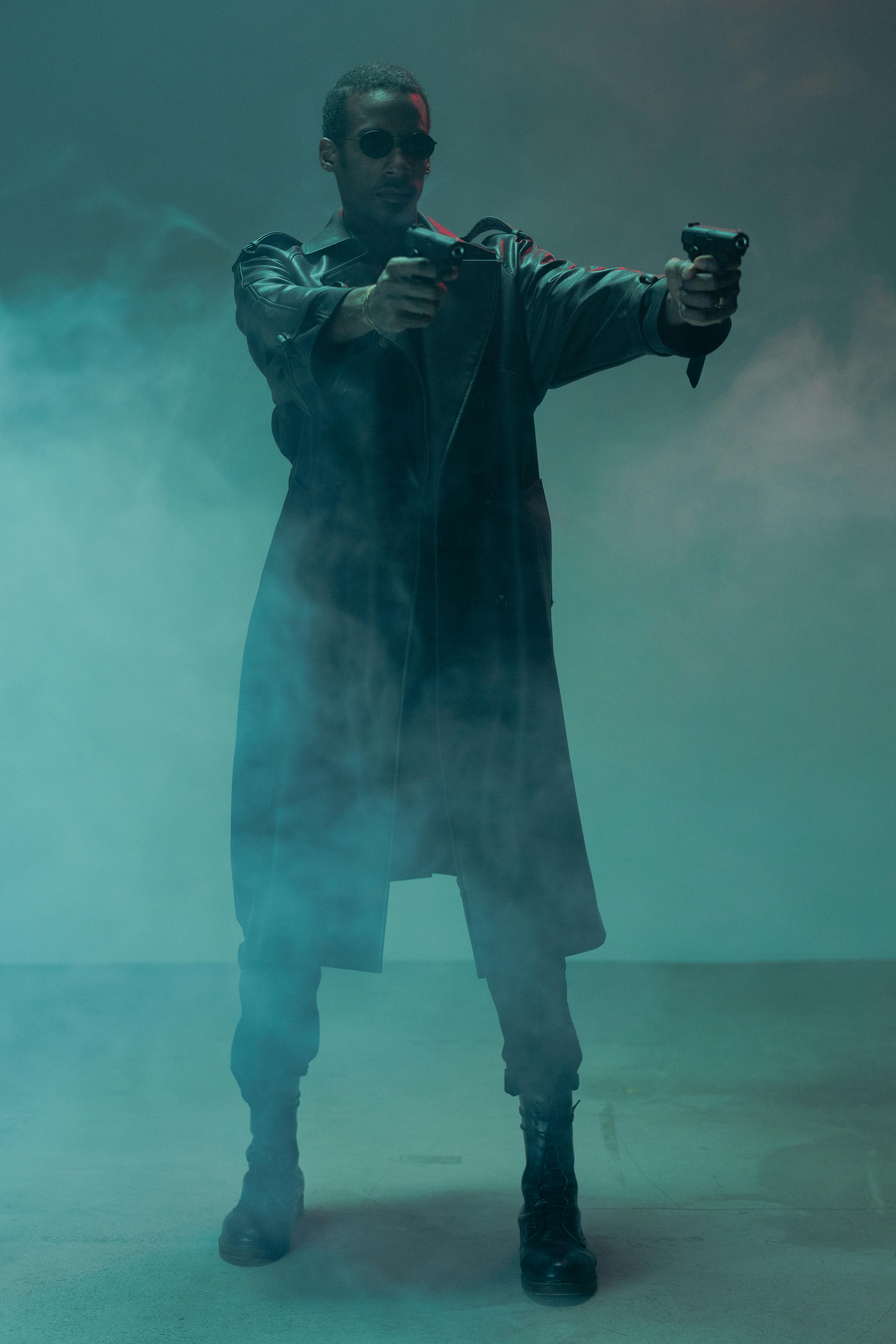 a fullbody shot of a man dressed in a long black leather coat and dark sunglasses, holding two pistols, stands in a smoky, dimly lit room with teal and blue tones.  the scene is dramatic and moody, evocative of film noir or cyberpunk aesthetics.  the man's expression is serious and intense.