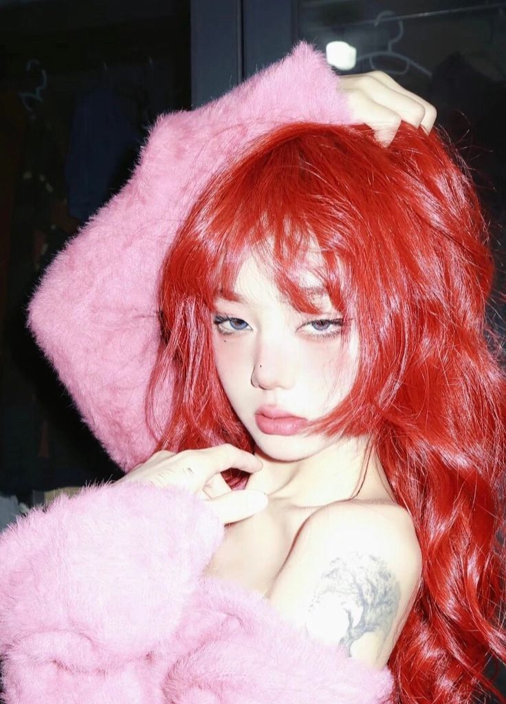 a young woman with vibrant red hair and a pink fuzzy sweater takes a selfie. she has trendy makeup with rosy cheeks and wears colored contact lenses. a floral tattoo is visible on her upper arm. the environment appears to be a clothing store with racks visible in the background. the lighting is bright and artificial, creating a slightly overexposed look.