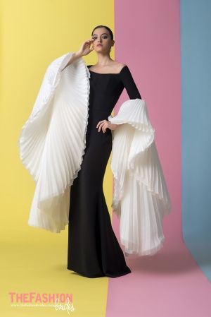 a woman in a black offshoulder gown with large white pleated sleeves stands in a studio setting. she is holding the dress with one hand and looking directly at the camera. the background is a combination of yellow, pink, and blue colors.