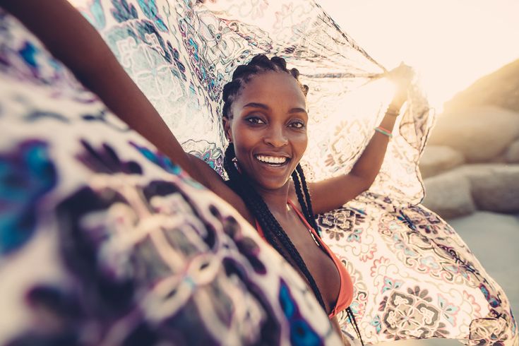 a black woman smiles brightly at the camera while holding a patterned sarong above her head. the sun shines brightly behind her, creating a backlit effect. the image is taken from a firstperson perspective, making the viewer feel as if they are standing directly in front of her.