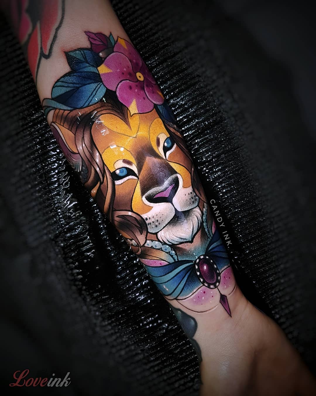 a neo traditional style tattoo of a lion's face with flowers on top and a bow tie on the bottom. the tattoo is on someone's inner forearm. the tattoo has a name on it in small lettering: "candy ink"