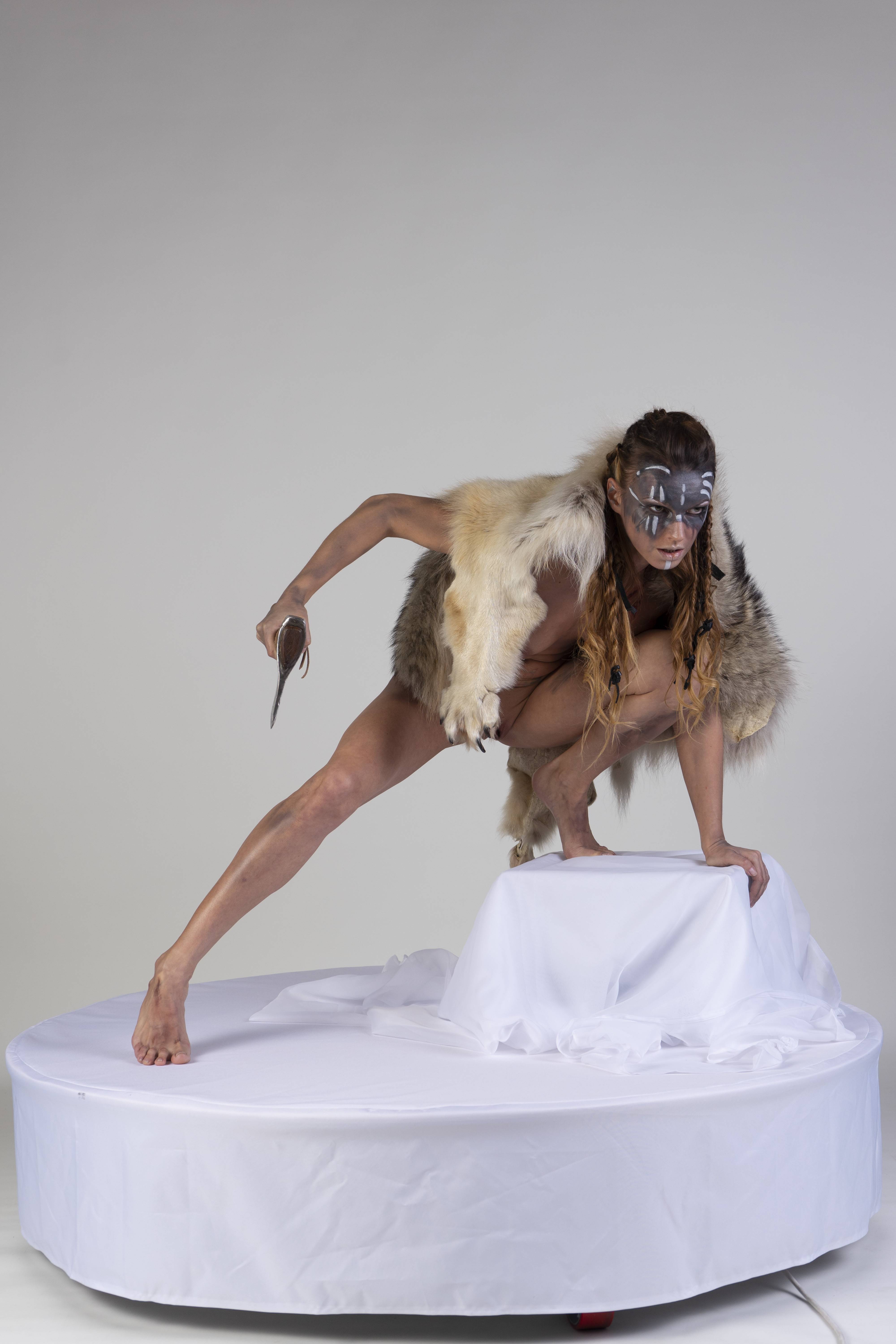 a woman with face paint, a fur vest, and holding a knife is crouched on a white platform against a white background. she is barefoot and appears to be posing or performing theatrically, with a tribal or warrior aesthetic.