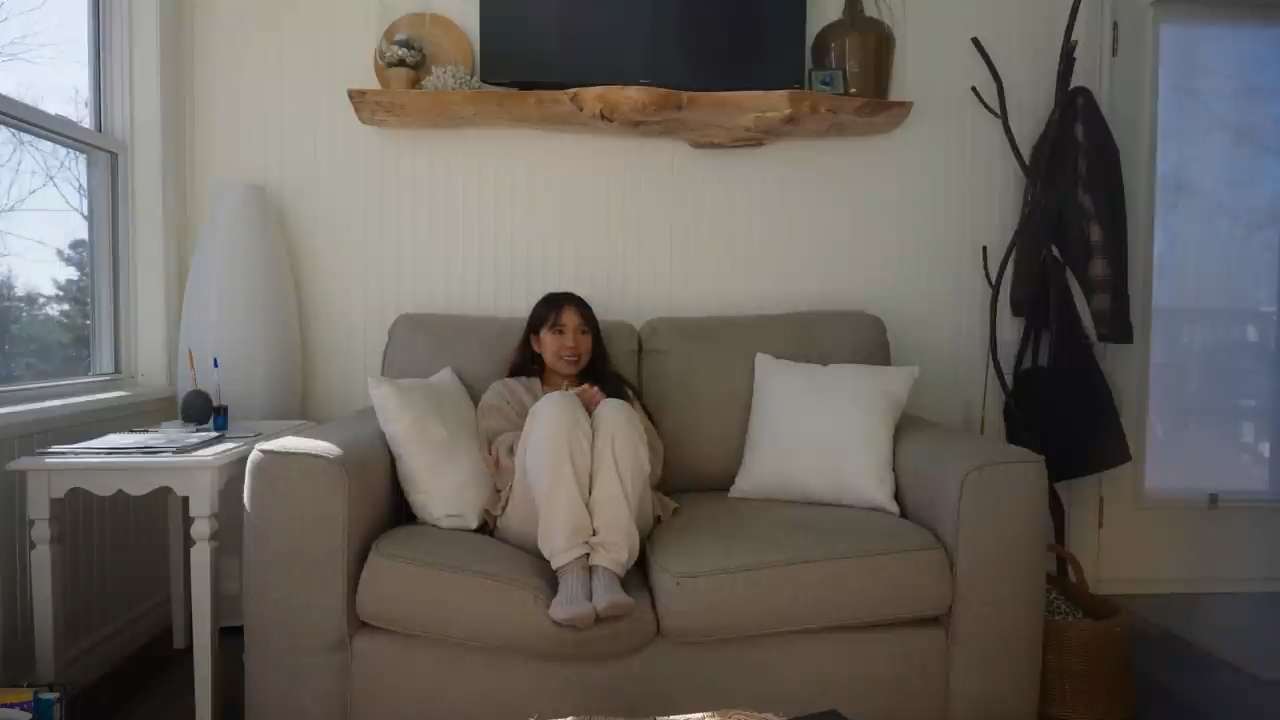 a young asian woman sits on a light beige couch in a naturally lit living room. she is wearing comfortable, lightcolored clothing and has a relaxed posture.  the room is minimally decorated with a wooden shelf above the couch, a floor lamp, and a small table beside the couch. a large window behind her brings in ample daylight, creating a bright and airy atmosphere. the overall feeling is one of peace and tranquility.