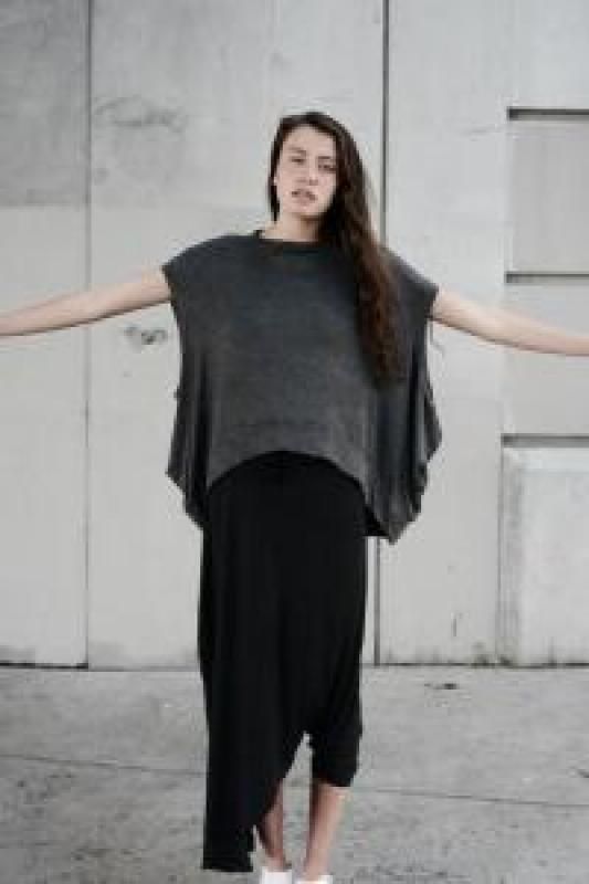 a woman stands in front of a concrete wall, wearing a casual outfit. she has a grey sleeveless top and a black asymmetrical dress. her arms are outstretched, and she is looking directly at the camera.