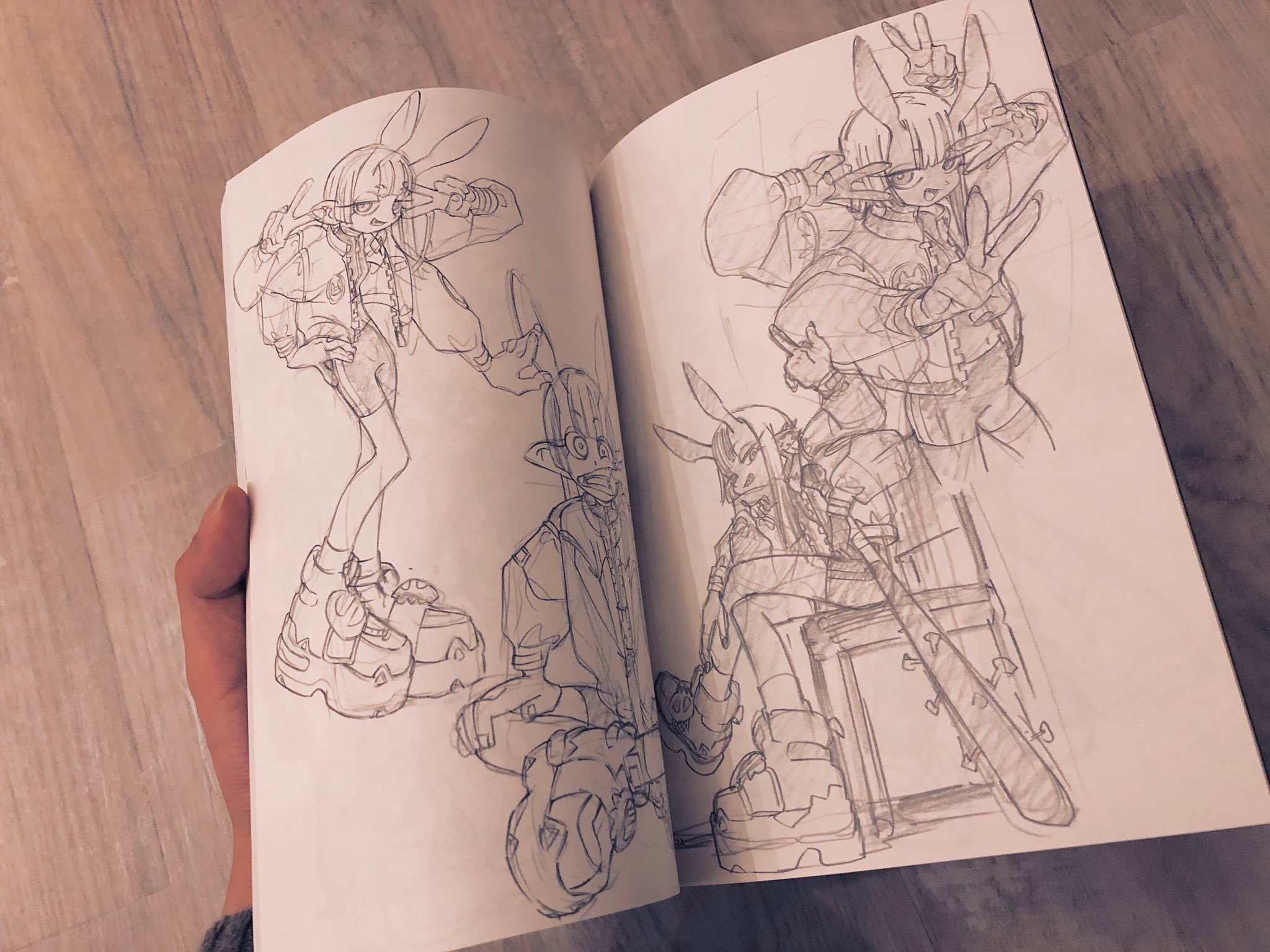 a hand holds open an artist's sketchbook displaying four pencil sketches of original characters. the characters all appear to be wearing some variation of  bunny ear hats, chunky platform shoes, and streetwear.  the sketches are unfinished and have been drawn with graphite pencils on offwhite paper. the background shows a light colored hardwood floor.