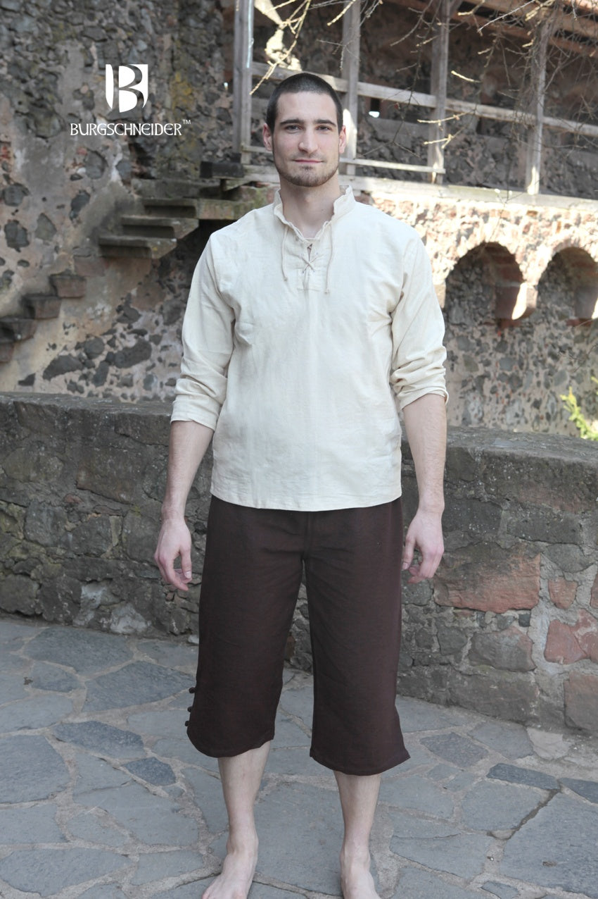 a man stands outdoors wearing medievalstyle clothing. he is wearing a lightcolored tunic with long sleeves and brown breeches. the man is barefoot and standing on a stone paved surface. behind him is a stone wall with a staircase and archways. the burgschneider logo is visible in the upper left corner of the image.