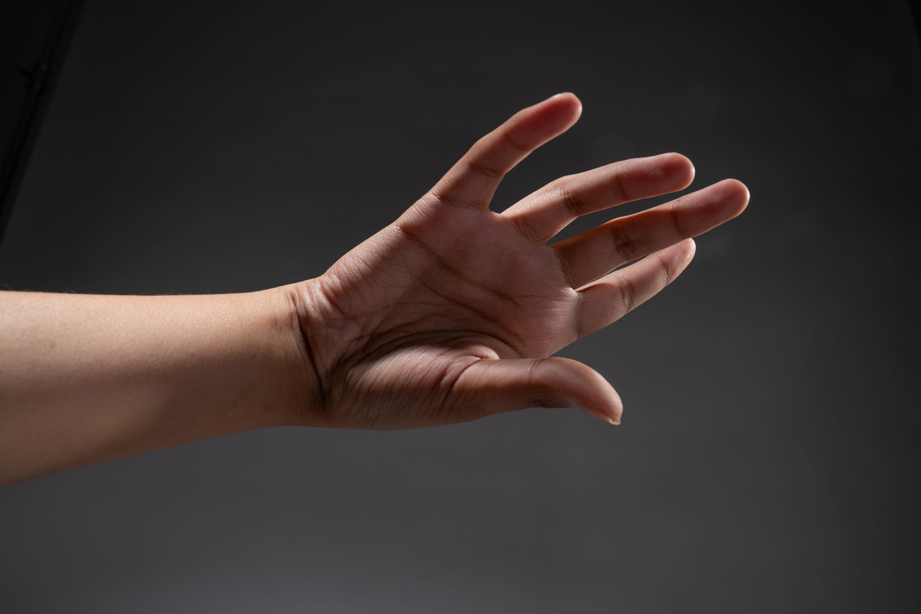 a hand is extended against a dark gray background. the hand is illuminated by a studio light, casting dramatic shadows. the fingers are spread apart in a gesture. the image focuses on the hand and arm, with no other elements in the frame.