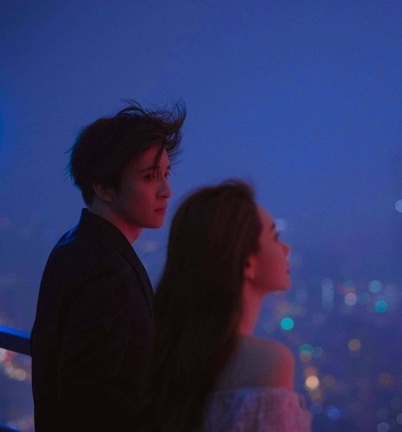 a man and a woman stand close together on a balcony at night, their backs to the camera as they gaze out at a cityscape. the scene is lit by dim, artificial light, casting a romantic neon glow.