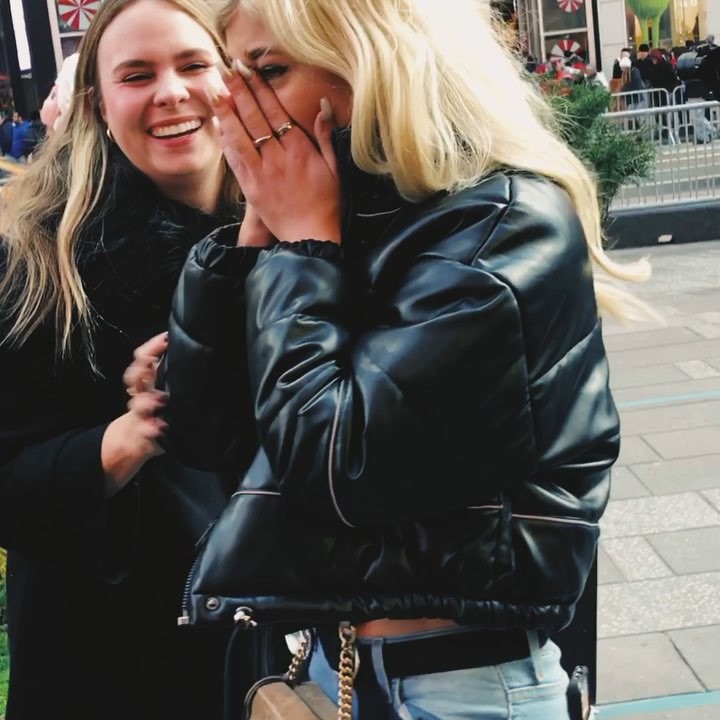 two women stand outside in a public space. one is laughing, while the other covers her face with her hands, wearing a black leather puffer jacket and denim jeans. in the background, there are other people and buildings.