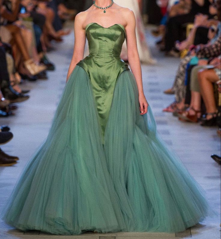 a woman wearing a green, strapless gown with a tulle skirt stands on a runway. there are people sitting on the sides watching. she is also wearing a green necklace.