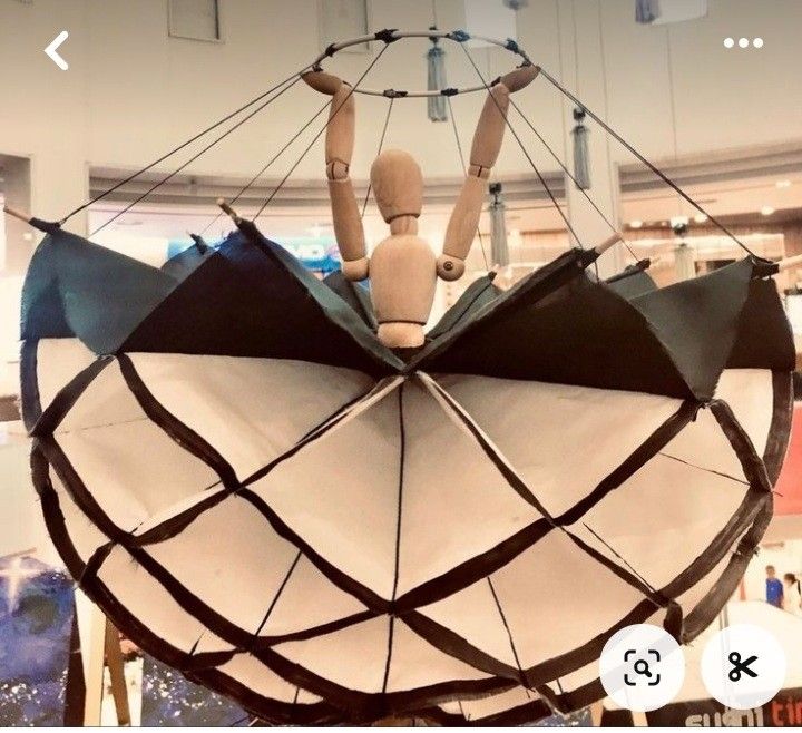 this image shows an artistic display hanging from the ceiling. it features a wooden mannequin figure with its arms raised, positioned in the center of an open umbrella. the umbrella is designed with alternating black and white panels and has a geometric pattern. the display is suspended from a circular frame using strings, creating an eyecatching and creative overhead installation.