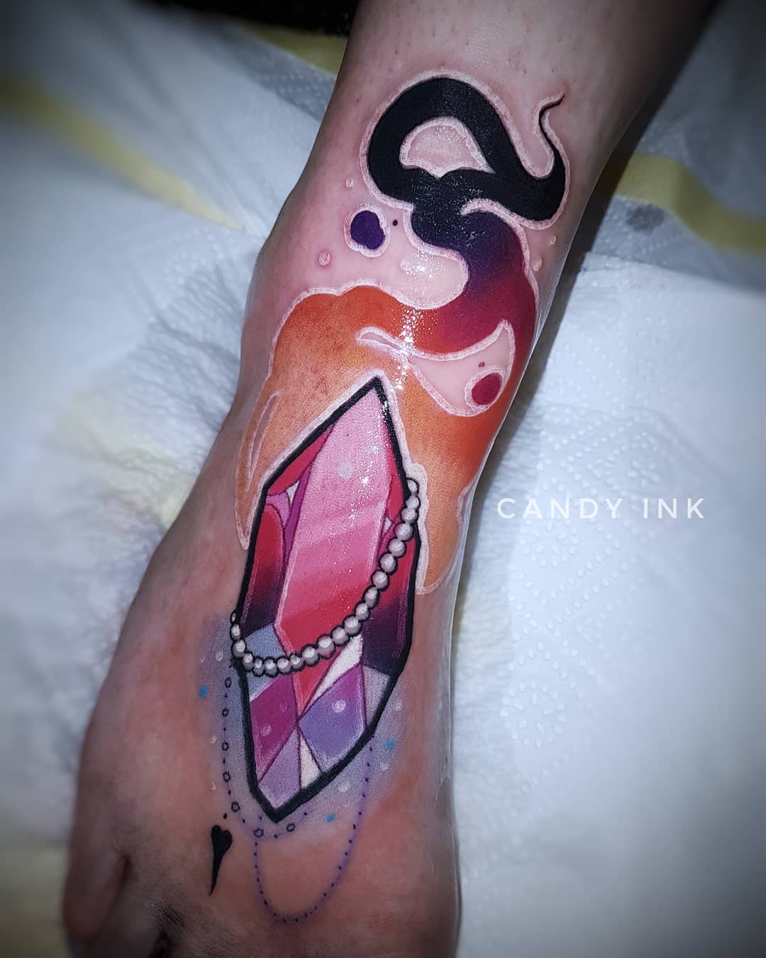 a person's foot with a tattoo of a pink and purple crystal with a beaded chain. the crystal is surrounded by yellow and orange flames and black smoke. the tattoo is done in a neotraditional style. the tattoo artist's watermark, "candy ink" is visible on the right side of the image.