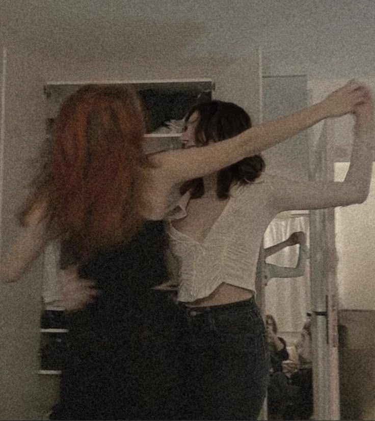 the image shows two women dancing in front of a mirror. one woman has red hair and is wearing a dark dress. the other woman has dark hair and is wearing a white shirt and jeans. they are standing in front of a wardrobe, and their reflections can be seen in the mirror. another person can be seen in the reflection of the mirror. the photo appears to be taken indoors.