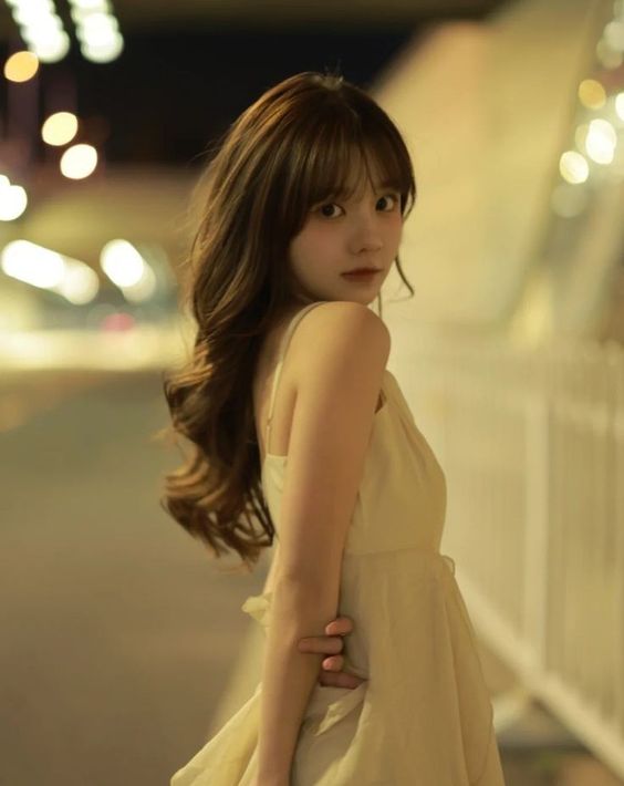 a young woman with long brown hair wearing a flowing white dress stands on a city street at night. she is looking over her shoulder at the camera, and the city lights create a warm, glowing background.