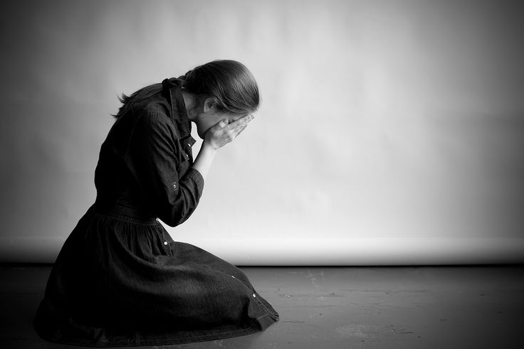 a woman in a long dress is kneeling on the floor with her face buried in her hands. she is crying and looks very sad. the image is in black and white, which adds to the sense of drama and melancholy.