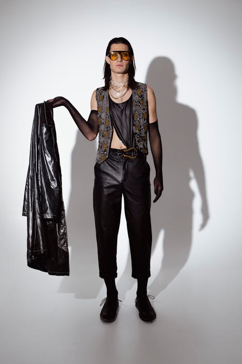 a full shot of a male model posing in a studio. the model is wearing a black leather jacket, black leather pants, a black vest with gold embellishments, black gloves, black boots, and yellowtinted sunglasses.  they also wear a layered necklace. the model has long dark hair. the background is plain white, casting a shadow of the model.  the model is holding a black leather jacket.