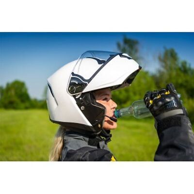 a woman wearing a white motorcycle helmet with a visor is drinking water from a bottle. she is wearing motorcycle riding gear and appears to be on a motorcycle, which is not shown in the photo. the background is a green field with trees in the distance.