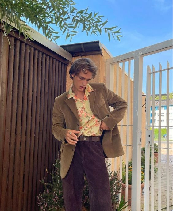 a young man wearing a brown jacket, light yellow floral shirt, brown corduroy pants and brown shoes stands in front of a white gate. the sky is blue and there are green plants and foliage around him.