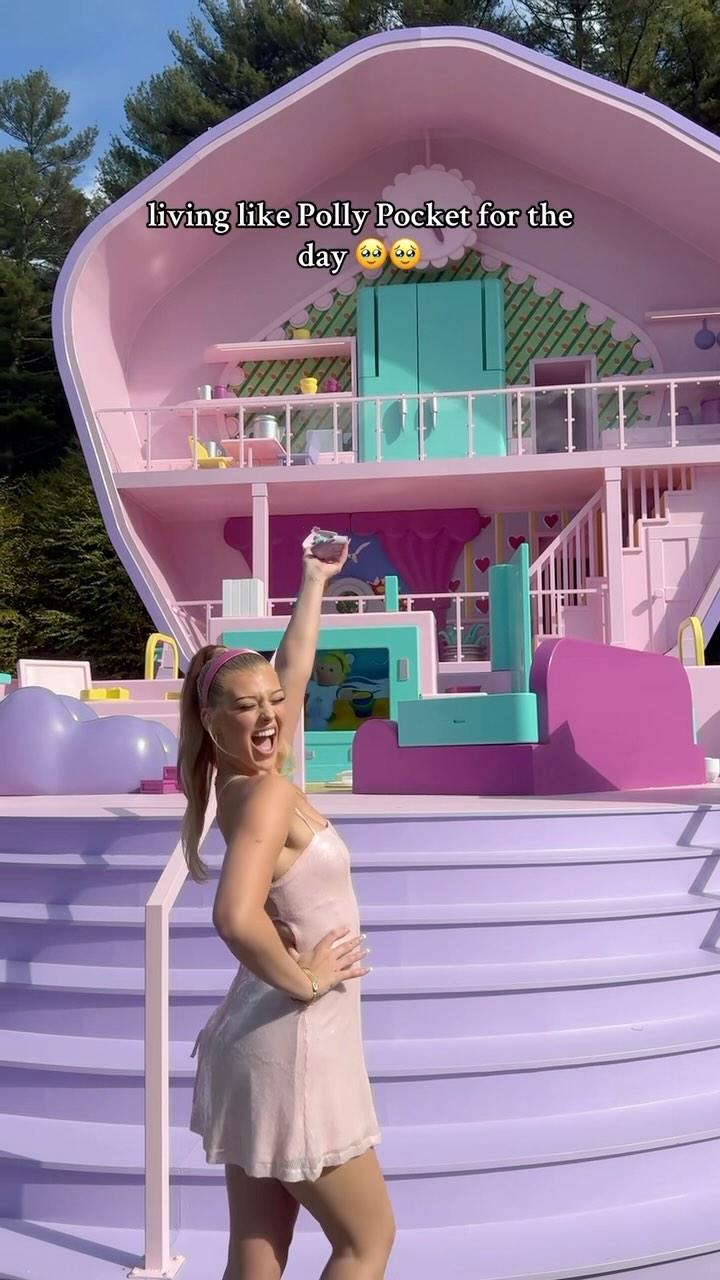 a woman is standing in front of a large, pink polly pocket dollhouse. she is wearing a light pink dress and a pink headband, and she is holding something in her raised right hand. the dollhouse has two floors and is surrounded by trees. the text "living like polly pocket for the day" is written on the image.