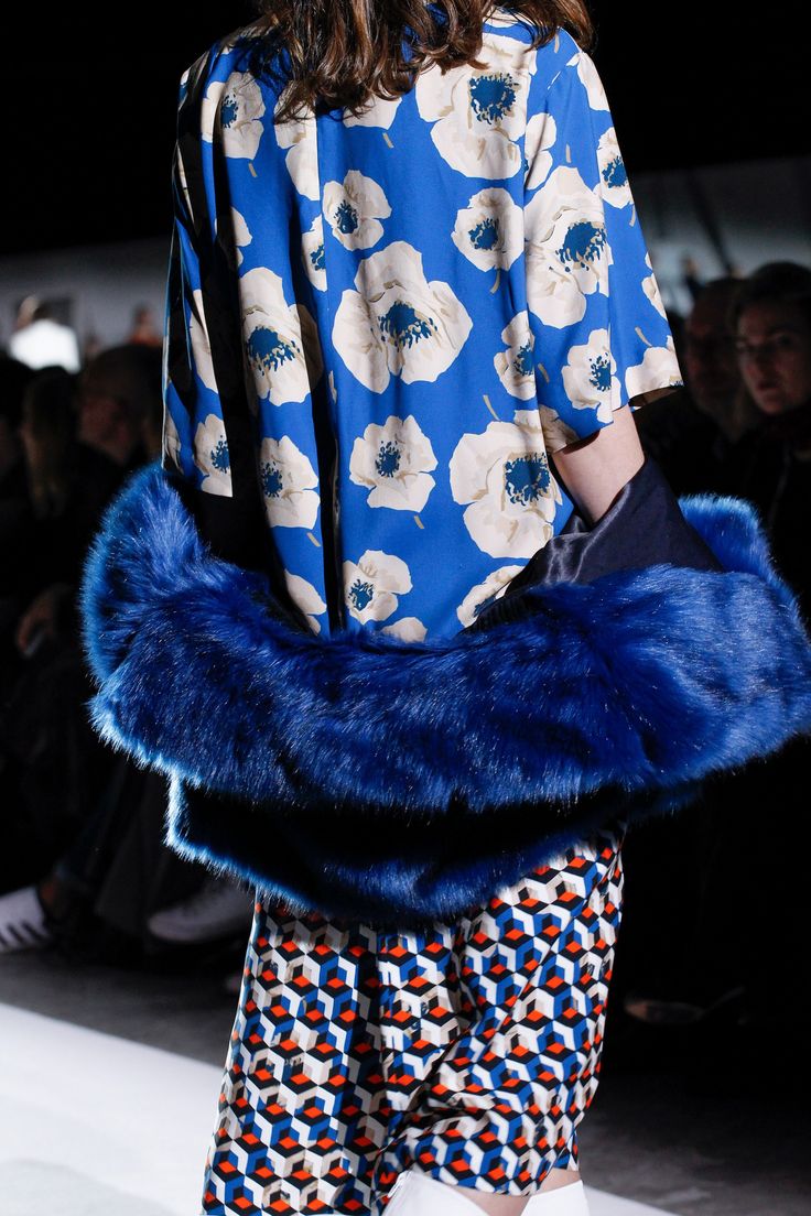 loseup view of a fashion model showcasing an outfit on a runway. the model is wearing a vibrant blue shirt with a white floral print. over this, she has a striking bold blue fur stole or wrap. the lower part of the outfit features a skirt with a geometric pattern in shades of orange, blue, and white. the overall style is bold, combining contrasting patterns and textures. the background is blurred, focusing the attention on the model's attire. the setting appears to be a fashion show or photoshoot.