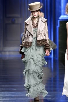 two female models walk the runway, the one in the front wearing a cream coat, hat, and a layered light blue ruffled dress. the model in the back is wearing a white dress. both women are posing for the cameras.